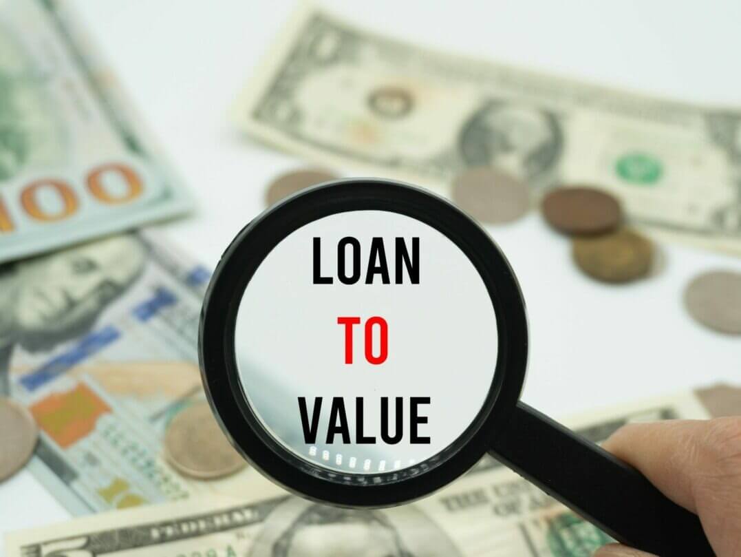 Magnifying glass focusing on the words "Loan to Value" with dollar bills and coins in the background.