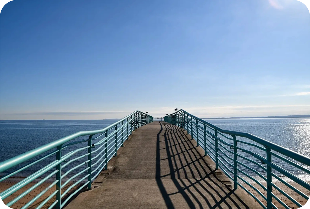 View down a curved boardwalk pier with benches and lamp posts overlooking the ocean