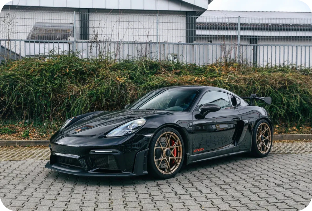Black Porsche 718 Cayman GT4 RS with bronze wheels and rear wing parked on a cobblestone driveway